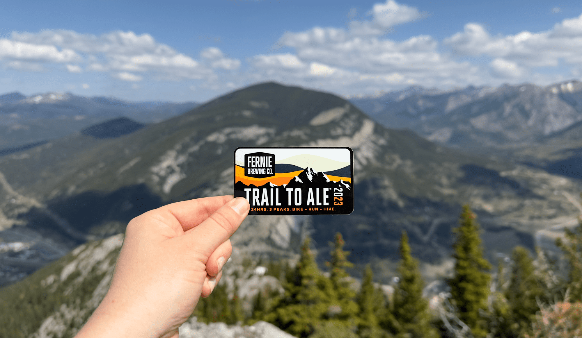 A photo of a person holding a 2023 Trail to Ale Sticker with a mountain in the background.
